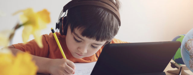 boy at desk with pencil and schoolwork wearing headphones with ipad in front of him