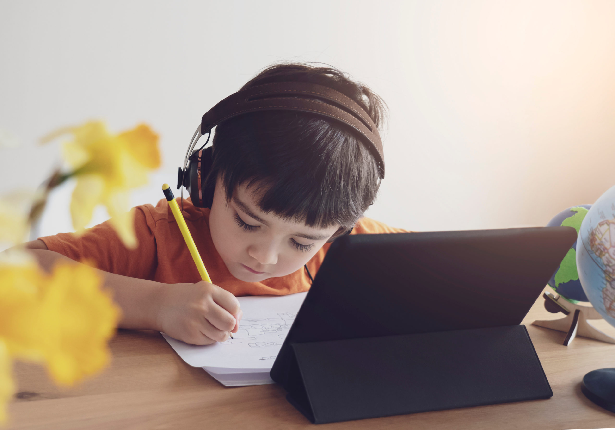 boy at desk with pencil and schoolwork wearing headphones with ipad in front of him