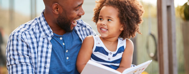 father sitting with daughter on lap and smiling at each other with a book