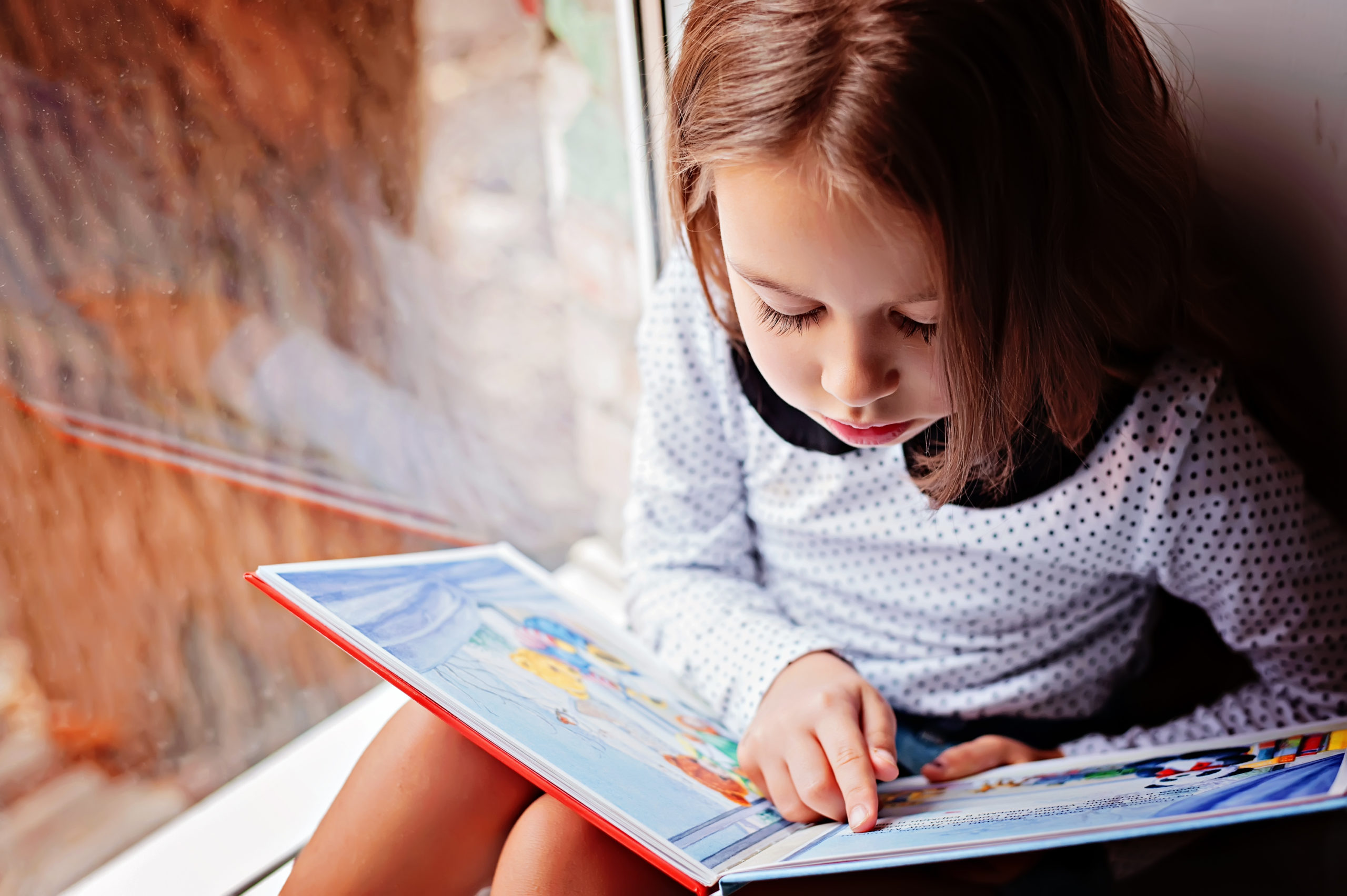 little girl reading book by window