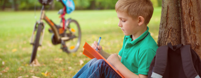 boy leaning against tree and writing with bicycle in background on the grass | summer learning ideas for kids