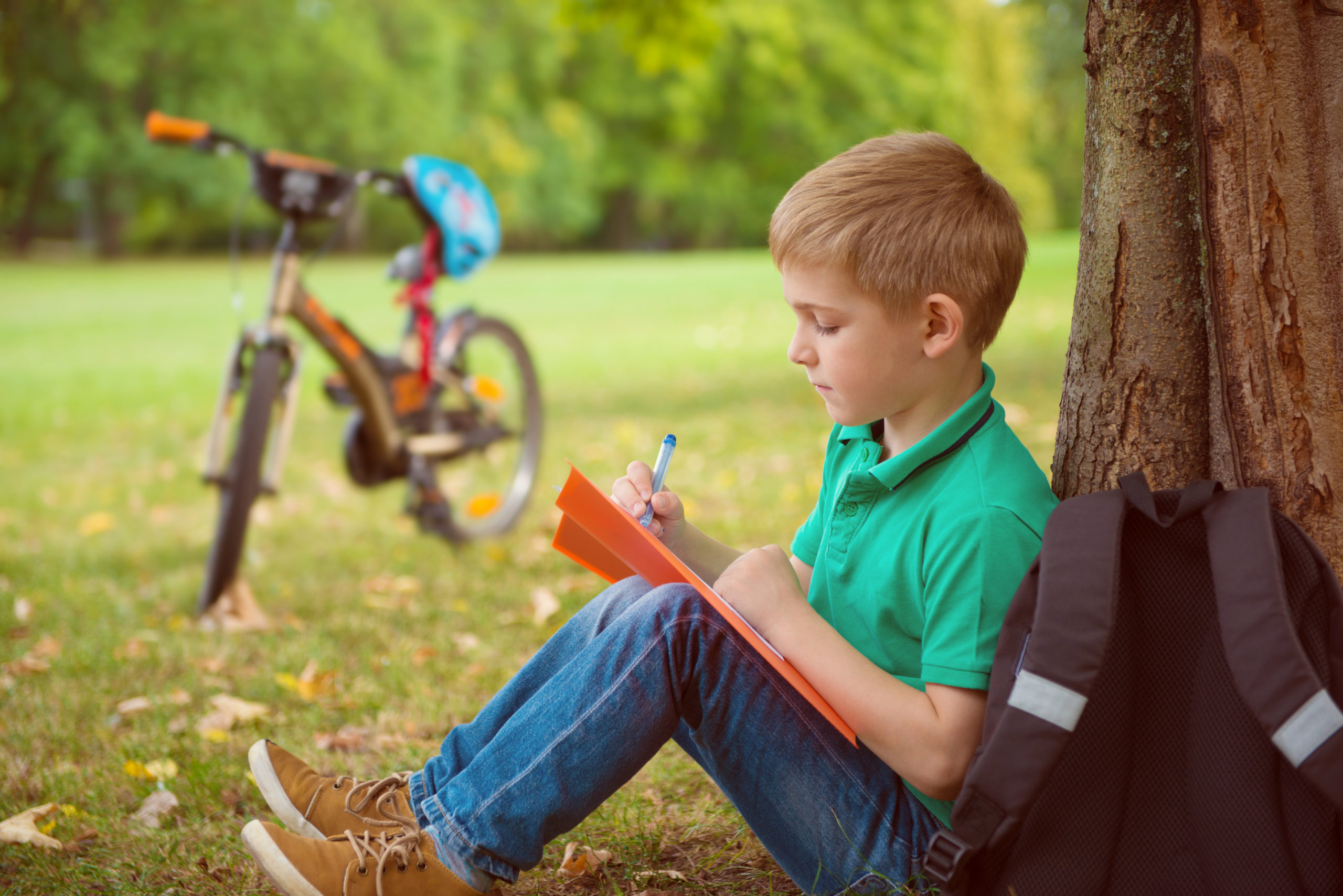 boy leaning against tree and writing with bicycle in background on the grass | summer learning ideas for kids