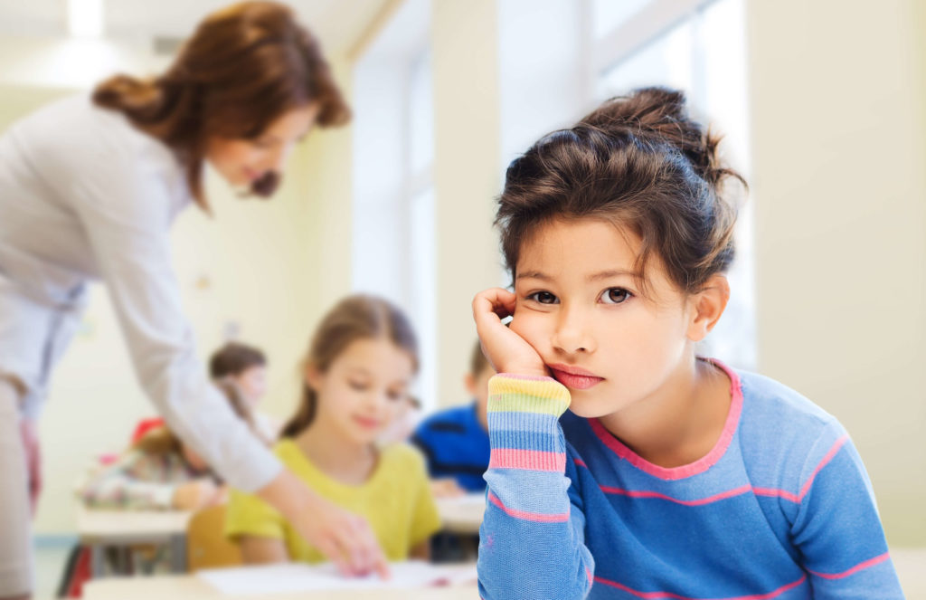 young female student sitting at desk looking bored with chin in hand, teacher helping other student in background